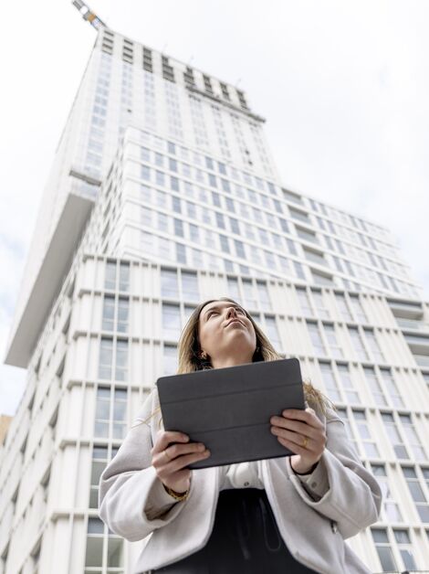 Blonde woman holding iPad looking up towards white apartment building.